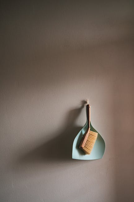 A light blue, teardrop-shaped wall hook mounted on a plain, smooth, beige-colored wall in a room with soft, natural lighting. Hanging from the hook is a small, rectangular wooden brush with light bris