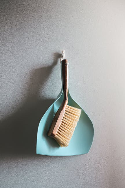 A light blue, teardrop-shaped wall hook mounted on a plain, smooth, beige-colored wall in a room with soft, natural lighting. Hanging from the hook is a small, rectangular wooden brush with light bris